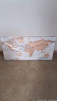 Full view of the horizontal rectangular wooden wall art showing all continents in natural wood tone on whitewashed wooden background, wrapped in plastic