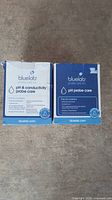 Two Bluelab branded boxes of probe care kits placed side by side on carpeted floor. Left box: pH & conductivity probe care kit. Right box: pH probe care kit.