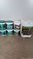 All plastic tubs and jug of organic fertilizers seen on floor in a row against white wall.