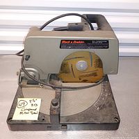 Side view photo of the Black & Decker compound miter saw sitting on its rotating table base. The saw blade is yellow and shows surface rust. Power cord wrapped partially around.