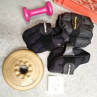 Black NordicTrack wraparound weights, Gold's Gym weighted glove, pink 2 lb dumbbell, and round gold 5 lb weight plate.