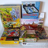 Contents of six jigsaw puzzle games including the Stow & Go roll-up mat, showing a variety of puzzle box designs generally featuring natural scenes with birds, flowers, hot air balloons, and tulip fields.