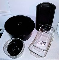 Black metal angel food cake pan, small clear glass fluted pan, two clear glass loaf pans with handles, and a black metal rectangular loaf pan displayed together on white surface