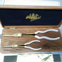 Two gold-plated long reach pliers with white handles in a wooden presentation box with black velvet lining.