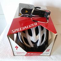 Box containing silver Specialized Chamonix adult helmet and Topeak bottle cage, showing packaging with Specialized branding and model information.