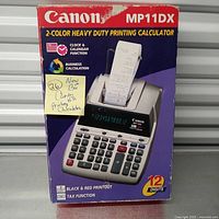 Photo of original Canon MP11DX packaging box with 2-color printing calculator image and feature highlights including clock & calendar function, business calculation, tax function, 12 digit display, black and red printout.