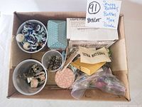 Top-down photo of a box containing various small containers and envelopes with sewing notions like buttons, bobbins, clips, beads, and threaders.
