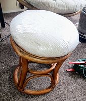 Front angled view of vintage rattan swivel ottoman with plastic covered wheat fabric cushion on pecan wood frame base.