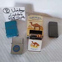 Photo showing four vintage lighters including one with a blue and gold emblem, one that resembles a Camel cigarette pack, one dark smooth metallic lighter, and a silver lighter with blue box.