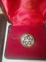 Close-up photo of the Millefiori glass tie tac in red-lined black jeweler's box showing colorful floral pattern and metal rope frame with chain.
