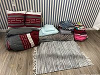 Photo of bedding items neatly folded and arranged on floor including comforter, pillows, weighted blanket, floor mats, and multiple sheets