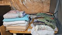 Stack of folded assorted bed blankets, house coat, and two cotton country-themed throws on a table in unfinished basement