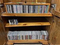 Wooden cabinet with two shelves holding CDs on the top and bottom shelves and cassette tapes on the middle shelf.