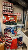Various Christmas decorations on a shelving unit including multiple unopened boxes of string lights, candy dishes with holiday figures, a Barbie stocking hanger, some stockings, and a candle holder.