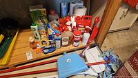Wide view of cleaning supplies, paper towels, broom, mop, and various bottles and containers on a wooden table in basement setting.