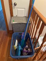 Grey fabric clothes hamper with hinged lid next to blue plastic laundry basket filled with cleaning supplies including Swiffer Wet Jet mop, Lysol wipes, broom, dustpans.