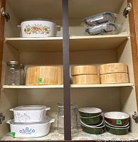 Wide view of cabinet shelf showing CorningWare casseroles, glass storage containers with lids, mason jars, wood salad bowls, small terrines, and milk glass ashtray.