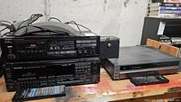 Three audio-video devices including Onkyo cassette deck stacked on Kenwood receiver and Mitsubishi DVD player on table with remotes
