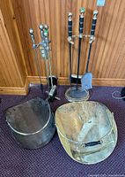 Two sets of fireplace tools with log caddies against wood panel wall on carpet floor