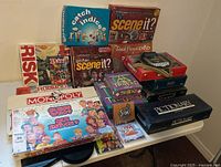 Wide shot of assorted board games and electronic games stacked on a white table, includes Risk, Monopoly, Scene It?, Guess Who?, Pictionary, and Sims games