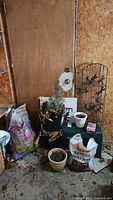 Overall gardening lot including plastic yard stool, bags of garden soil, planter pots and metal trellises leaning against a wooden wall inside a garage.