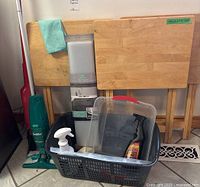 Three wooden TV tables, broom, Swiffer mop, laundry basket with spray bottle and containers, and a new under sink mat visible.