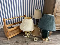 Photo showing two matching table lamps with beige pleated shades, a wooden magazine rack, a desk lamp with green ceramic base and blue-green shade, a smaller desk lamp with carved decorative base and beige shade, power bar, and vintage alarm clock on floor against striped wallpaper.