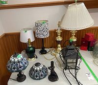 Eight lamps on a white table and floor, including two Tiffany style stained glass lamps, a hanging lantern, and various bases and shades such as ceramic green, brass, and black carved base lamps with fabric and paper shades.