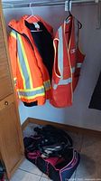Image showing orange work jacket with retroreflective stripes and orange reflective vest hanging next to each other, with black and pink bag on the floor beneath.