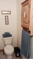 Bathroom corner showing pine cabinet above toilet, blue towels folded on toilet tank, black waste basket on floor.