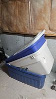 Two stacked plastic storage totes: a beige tote with a blue mismatched lid on top of a blue tote in basement location with concrete floor and unfinished walls.