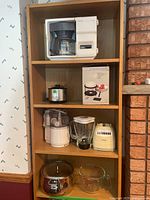 Wood shelving unit showing all items in lot including coffee maker, crockpot, fondue set, food processor, blender base, sauce pot, and measuring cup.