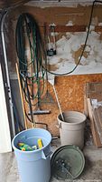 Garage corner with two garbage pails, various shovels, rake, axe, brooms and garden tools leaning against wall with a hanging green garden hose.