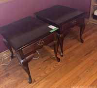 Two dark stained wooden side tables with curved legs and brass handles on the drawers, positioned on a hardwood floor near a purple wall.