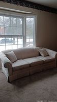 Full view of the sofa in front of large window, showing 3-seater beige fabric, two pillows