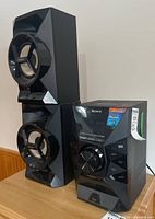 Sony home audio system with two black two-way speakers, angled geometric design with circular drivers and central unit with display and control buttons.
