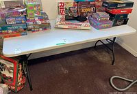 Wide shot of the white folding plastic utility table with various boxed games on top, showing the overall structure and condition.