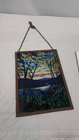 Photo of scenic stained glass wall hanging depicting trees, mountains, water, and sky at sunset with white flowers and foliage.