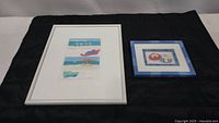 Two framed prints placed side by side, one large rectangular white framed print with beach scenes, one smaller square blue framed print with water baby images.