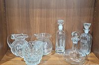 Photo of the set showing three glass pitchers, three glass decanters with stoppers including etched and vintage designs, on wooden shelf.