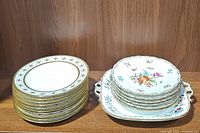 Stack of 10 Limoges gold-trimmed plates and 6 Dresden floral plates on a decorative tray, displayed on wooden shelf.