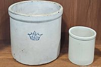 Both stoneware crocks together on wooden shelf against brown backdrop. The larger crock has a blue crown and number 2 logo; the smaller is plain.