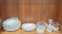 Eight assorted cut crystal items displayed on a wooden shelf including bowls, candy dishes, pitcher, vase, and lidded jelly jar.
