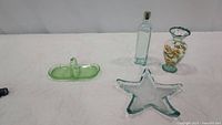 Four decorative glass pieces on a white cloth background including uranium glass handled dish, starfish star-shaped candy dish, painted glass vase, and tall green glass jar with cork stopper.