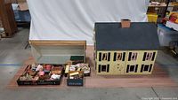 Front view of handcrafted yellow doll house with windows and black shutters. Next to it are two trays filled with miniature furniture and accessories.