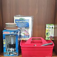 Lot contents showing all items: Coleman LED lantern in box, red tool bin with tools including hammer, Garden Harvest-Guard protective cover, plant saucers stacked, vintage metal utensil windchime