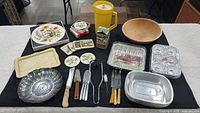 Full lot overview on black cloth: vintage Tupperware pitcher (yellow), wooden serving bowl, ceramic trays and plates, assorted kitchen knives and utensils, new foil pans, place mats, coasters