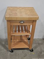 Front view of the butcher block kitchen cart with drawer closed, showing the thick wooden top, drawer handle, and two shelves below on caster wheels.