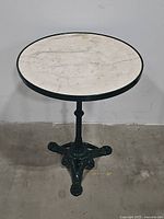 Full view of the round marble-top bistro table with black metal rim and ornate black tripod pedestal base