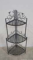 Front view of the 3-tier black metal corner shelf showing lattice shelving and bird on branch design.